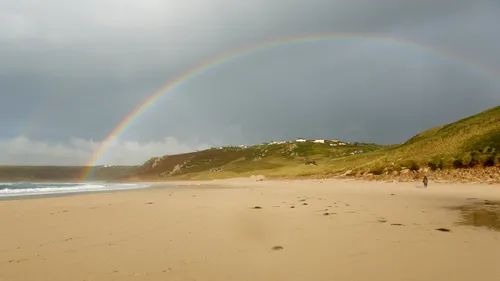 Sennen rainbow This landscape photograph shows the coast at Sennen Cove during a late afternoon in early autumn. The main subject of the image is the Sennen rainbow, which forms a distinct arc across a sandy beach in front of grassy hillsides. In the distance, houses are scattered on the hillside above the cove. The sky is partly cloudy and the rainbow's colours are vivid, adding a striking feature to the scene. The coastline of Sennen Cove and the stretch of beach are clearly visible, with gentle waves approaching the shore. The image captures the natural features of the Cornish coast in autumn, emphasising the beach, the landscape and the presence of a rainbow above the cove.